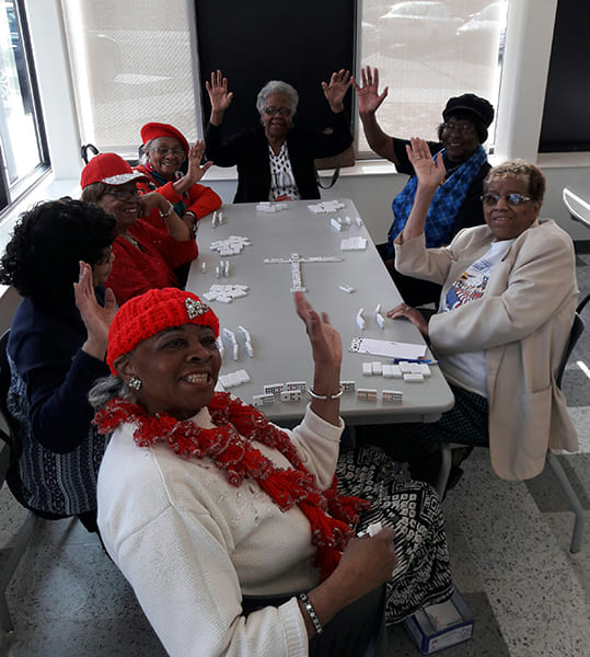 Seven Rose Centers participants sitting at a table playing dominoes while smiling and waving